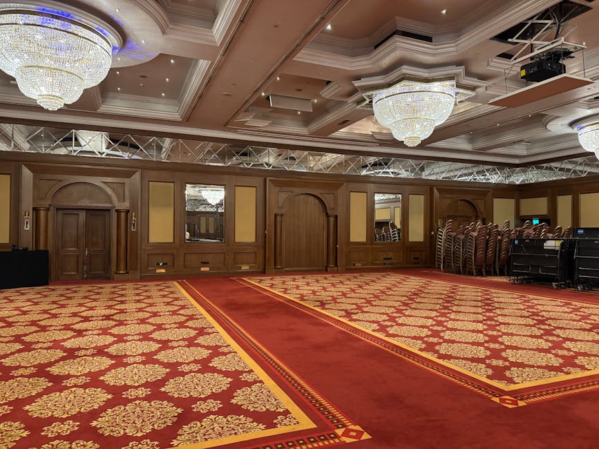 A large, ornate conference room featuring a plush red carpet with intricate gold floral patterns, illuminated by grand crystal chandeliers hanging from the decorated ceiling. The room has wood-paneled